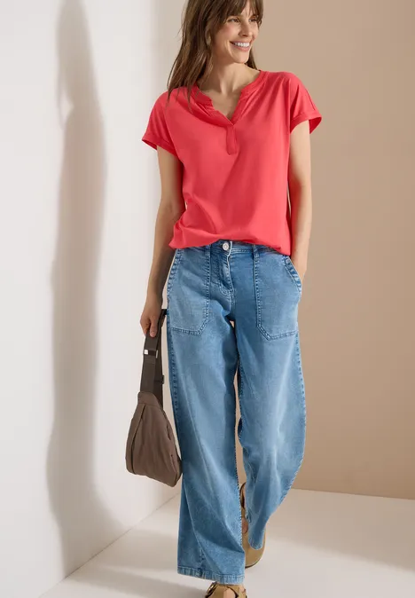 Kurzarm Shirt im Tunika-Look coral Frau in rotem T-Shirt, weiten Jeans und brauner Handtasche vor neutralem Hintergrund.
