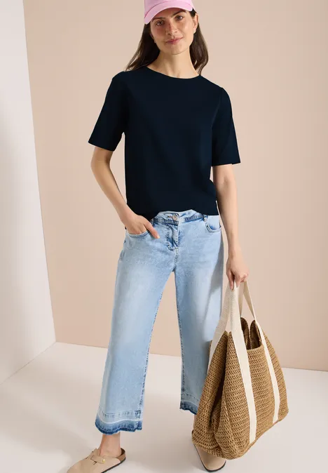 Basic Shirt met boothals universal blue Vrouw in zwart t-shirt, lichtblauwe wijde jeans en strooien tas voor een lichte achtergrond.