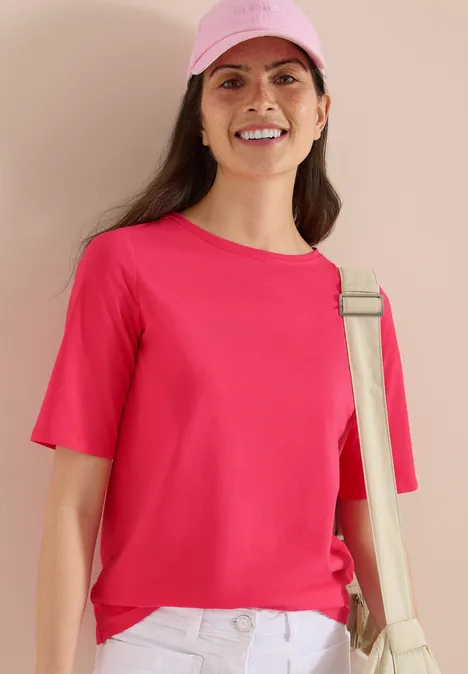 Basic Shirt met boothals geranium red Vrouw met roze T-shirt, witte broek en beige schoudertas, roze pet, neutrale achtergrond.