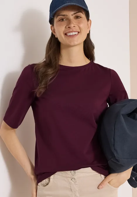 Basic Shirt met boothals sweet grape red Vrouw met bordeauxrood shirt en pet, beige broek, houdt donkerblauwe jas vast voor neutrale achtergrond