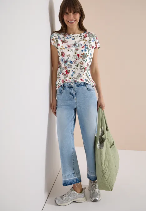 Shirt met bloemenprint light alabaster white Vrouw in wit bloemen-T-shirt, lichtblauwe jeans, zilveren sneakers en groene gewatteerde tas.