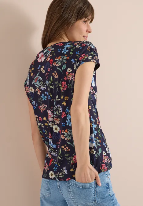 Shirt met bloemenprint universal blue Een vrouw draagt een kort donkerblauw shirt met kleurrijk bloemenpatroon en een spijkerbroek, zijaanzicht.