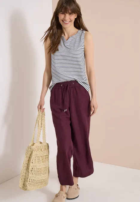 Dame trägt gestreiftes ärmelloses Top, weite violette Leinenhose und Basttasche.