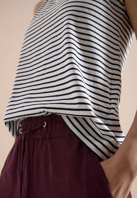 Gestreiftes Oberteil in Weiß und Dunkelblau, bordeauxrote Hose mit Tunnelzug und Hand in Tasche.