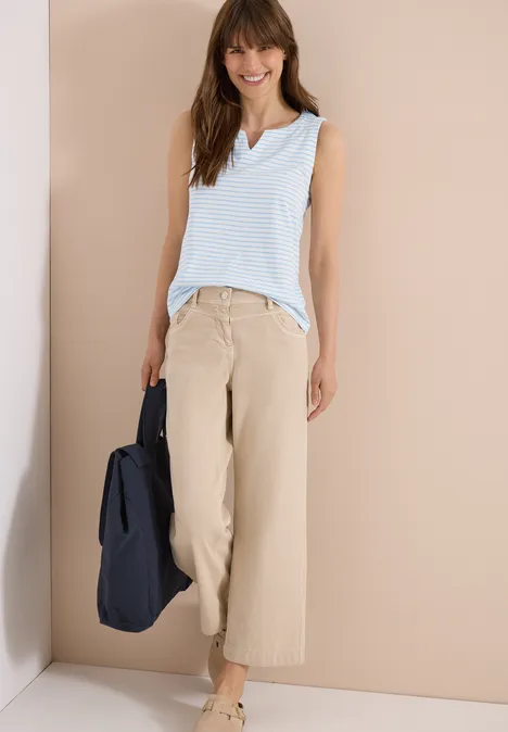 Frau in gestreiftem hellblauen Top und beigen weiten Hosen, trägt eine dunkelblaue Tasche, steht vor hellem Hintergrund.
