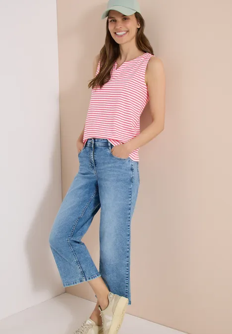 Eine Frau lehnt an einer Wand und trägt ein pink-weiß gestreiftes Top, weite Jeans und Sneaker.