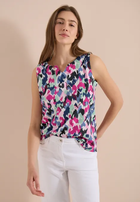 Vrouw draagt kleurrijke mouwloze top met abstract patroon en witte broek voor neutrale achtergrond.