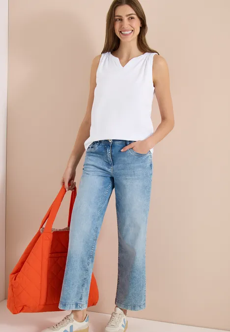 Frau in weißem Top und hellblauen Jeans mit großer orangefarbener Tasche vor neutralem Hintergrund.