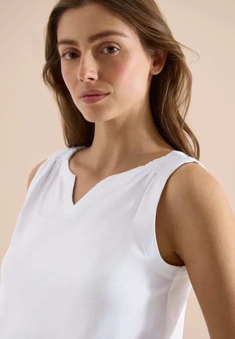 Frau in weißem, ärmellosem Top mit V-Ausschnitt, die entspannt posiert.