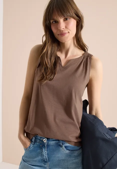 Frau in braunem, ärmellosem Top und Jeans, hält eine Jacke in der Hand.