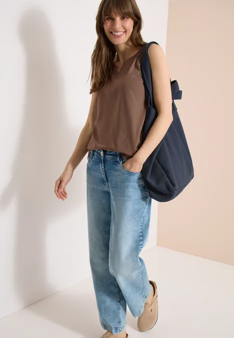 Eine lässige Person trägt braunes Top, weite Jeans, beige Schuhe und eine dunkelblaue Tasche vor hellem Hintergrund.
