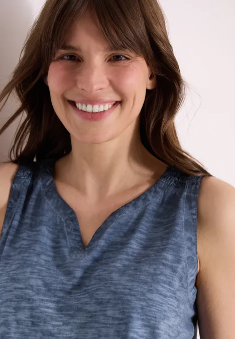 Eine Person trägt ein ärmelloses, blaues Oberteil mit V-Ausschnitt vor hellem Hintergrund.