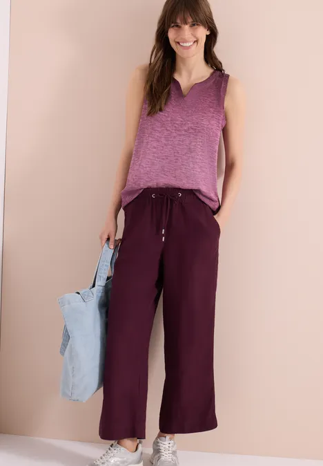 Frau in lila ärmellosem Top, weiten weinroten Hosen und hellblauer Tasche vor neutralem Hintergrund.