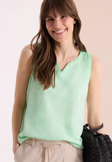 Eine Frau trägt ein ärmelloses mintgrünes Top, helle Leinenhosen und hält einen handgewebten Korb.