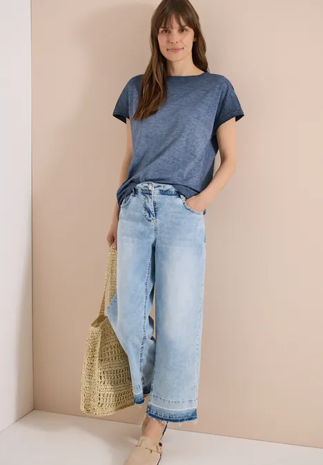 Frau lehnt an beiger Wand, trägt blaues T-Shirt, weite Jeans und hält eine geflochtene Tasche.