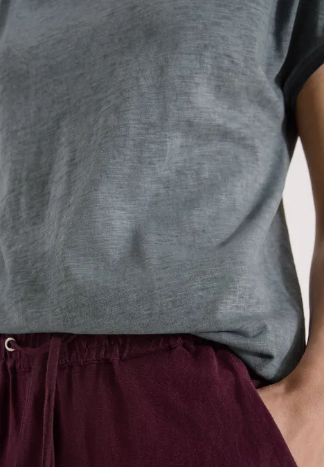 Oberkörper mit grauem T-Shirt und bordeauxroter Hose, Person steht entspannt mit Hand in Tasche.