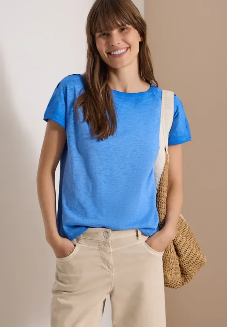 Shirt van geverfde jersey surf blue Vrouw in blauw T-shirt, beige broek en strooien tas poseert voor een effen lichte achtergrond.