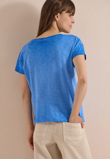 Shirt van geverfde jersey surf blue Vrouw van achteren met blauw shirt en beige broek, met één hand in de zak.