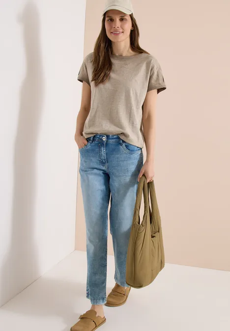 Frau trägt beige T-Shirt, blaue Jeans und braune Hausschuhe, hält olivgrüne Stofftasche.