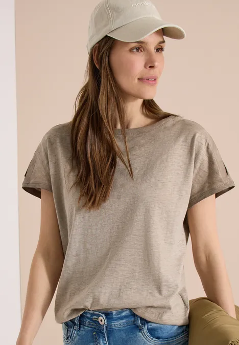 Frau mit Baseballcap, grauem T-Shirt und Jeans hält eine grüne Tasche vor neutralem Hintergrund.