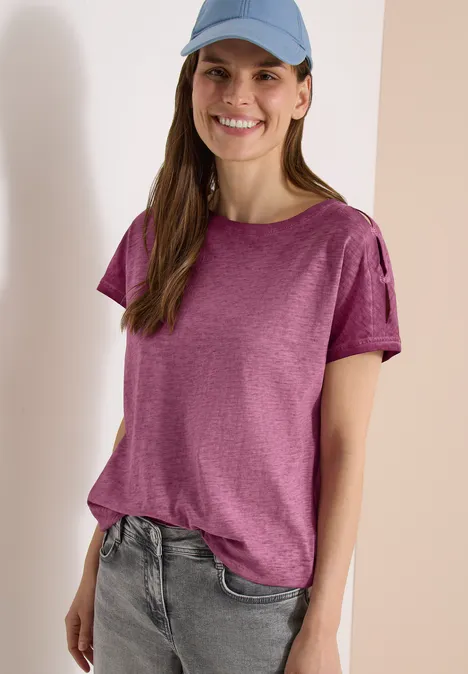 Eine Frau in lässigem Outfit: violettes T-Shirt, helle Jeans und blaue Cap vor hellem Hintergrund.
