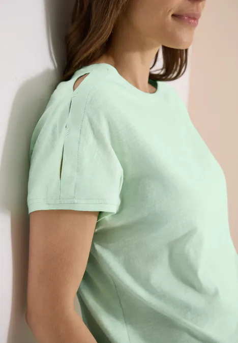 Seitliche Ansicht einer Frau in mintgrünem Kurzarmshirt mit Schlitzdetail am Ärmel, an hellem Hintergrund.