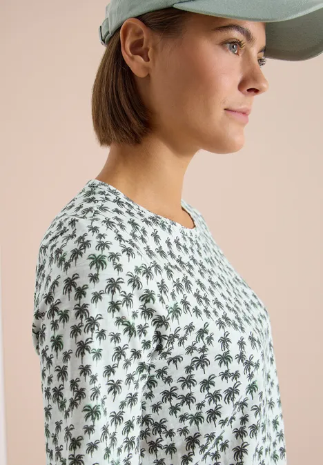 Seitliche Ansicht einer Frau mit Cap, die ein weißes T-Shirt mit grünen Palmenmuster trägt.