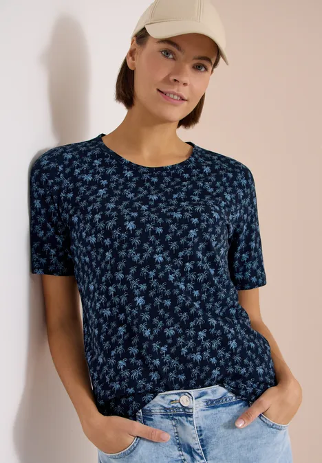 Frau lehnt an Wand, trägt dunkelblaues, geblümtes T-Shirt, helle Jeans und Cap, Hände in Taschen.