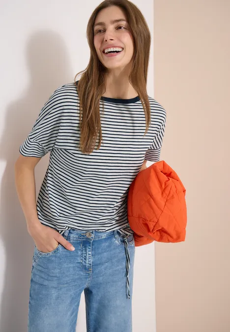 Frau in gestreiftem Shirt und Jeans hält eine orangefarbene Jacke locker in der Hand.