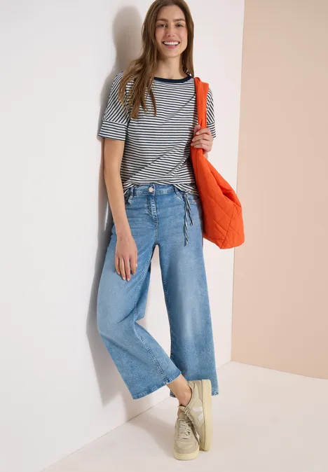 Frau lehnt an weißer Wand, trägt gestreiftes Shirt, weite Jeans, rote Tasche und beige Sneaker.