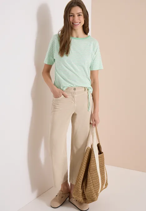 Frau lehnt an Wand, trägt mintgrünes T-Shirt, helle Stoffhose, Basttasche und flache Schuhe.