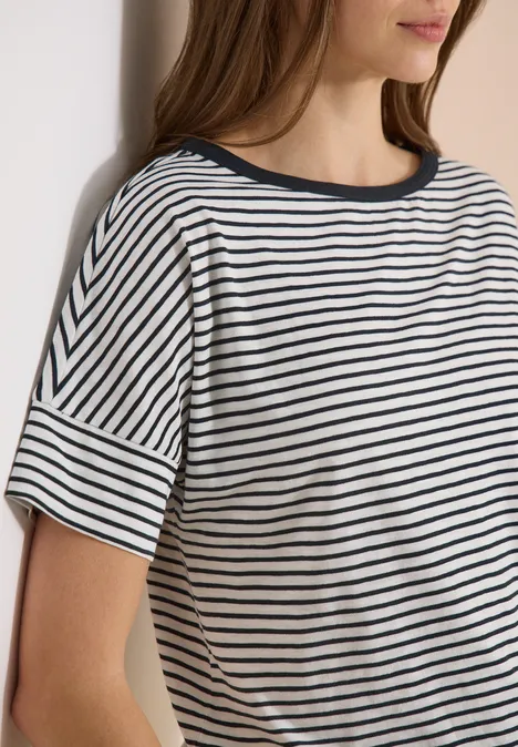 Frau trägt ein weiß-navy gestreiftes T-Shirt mit kurzen Ärmeln und lehnt an einer Wand.