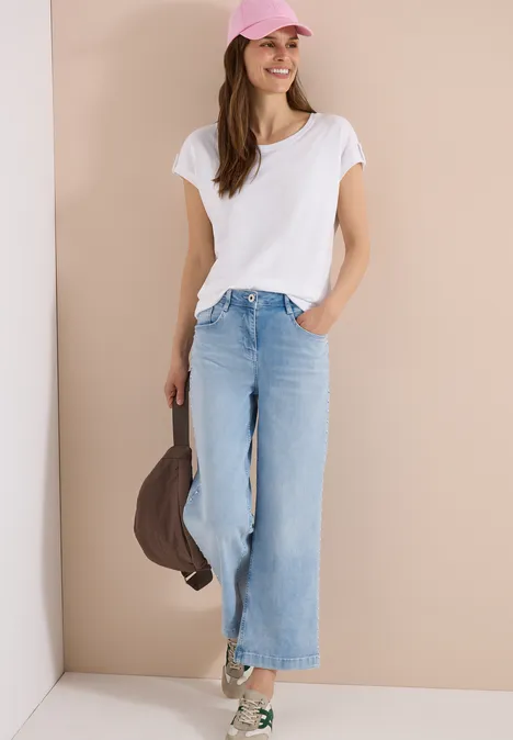 Frau in weißem T-Shirt, weiten hellblauen Jeans und Baseballcap lehnt an Wand, hält Jacke