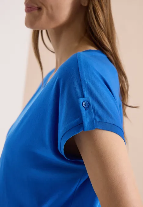 Zijaanzicht van een vrouw in een blauw t-shirt met korte mouwen en knoopdetail.
