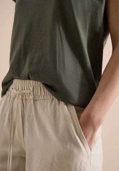 Grünes ärmelloses Top mit beiger Hose, Hand entspannt in Hosentasche.