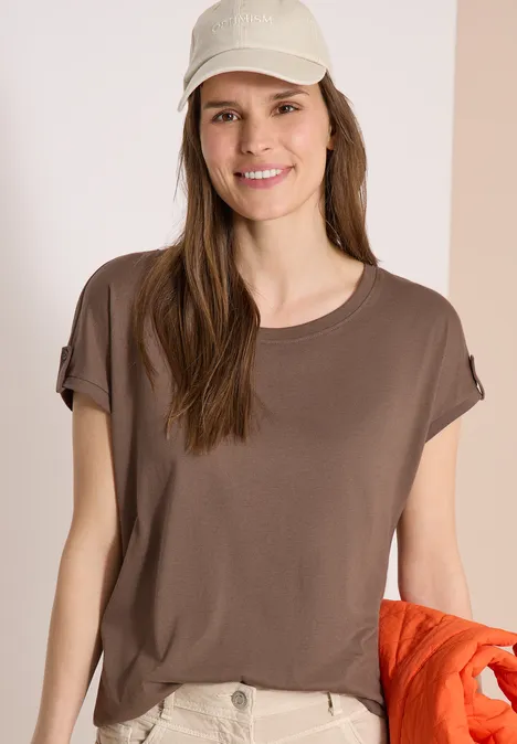 Vrouw in bruin T-shirt en beige broek houdt een oranje jasje vast tegen lichte achtergrond.