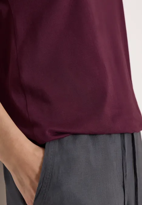 Bordeauxfarbenes Oberteil, graue Hose, eine Hand steckt in der Tasche.