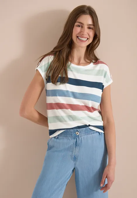 Een vrouw poseert voor een neutrale achtergrond, met een gestreept T-shirt en lichte spijkerbroek.