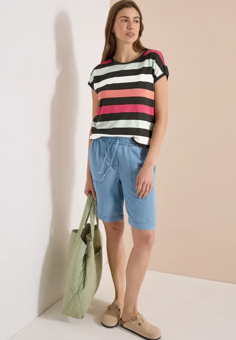 Une femme en t-shirt rayé multicolore, bermudas bleu clair, sac vert et mocassins.