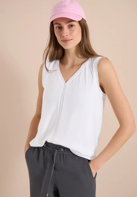 Frau in weißem ärmellosen Top und grauer Hose, Hände in den Taschen.