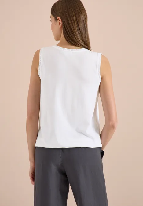 Frau steht mit dem Rücken zur Kamera, trägt ärmelloses weißes Top und graue Hose.