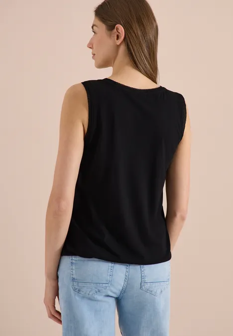 Vrouw van achteren in een zwart mouwloos shirt en lichtblauwe jeans voor een effen achtergrond.