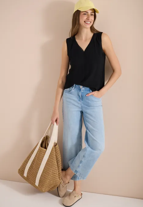 Vrouw in zwart mouwloos topje, lichtblauwe jeans, loafers, met een strooien tas voor een beige achtergrond.