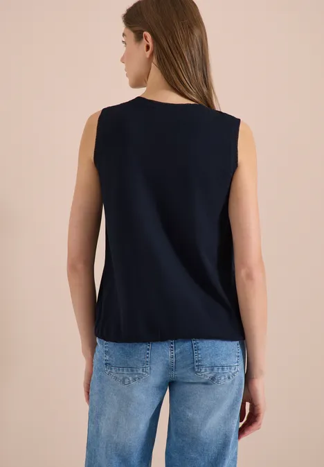 Achteraanzicht van een vrouw in een mouwloos donkerblauw topje en lichtblauwe spijkerbroek tegen neutrale achtergrond.