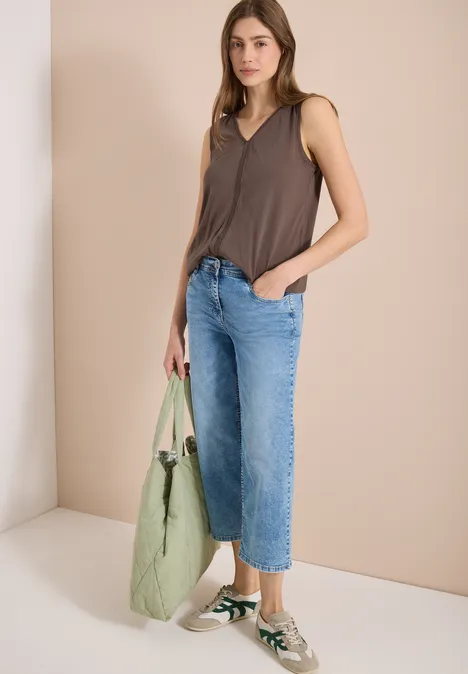 Vrouw met bruin topje, losse jeans, groene tas en sneakers voor een neutrale, lichte achtergrond.
