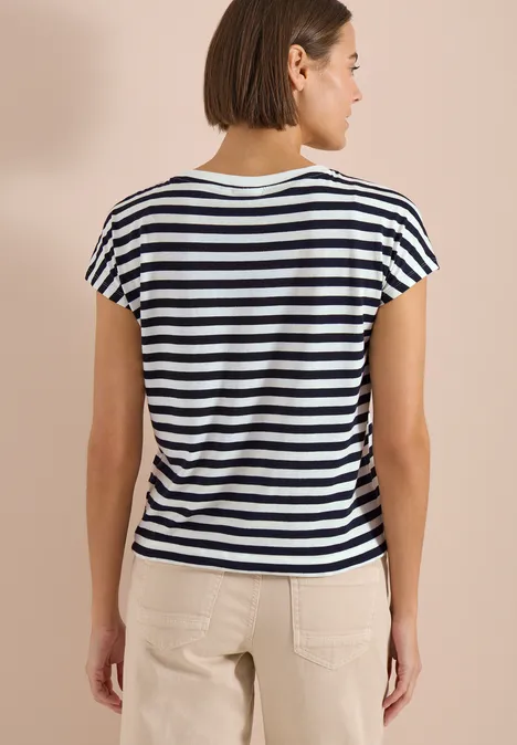 Une femme de dos porte un tee-shirt rayé noir et blanc à manches courtes et un pantalon clair.