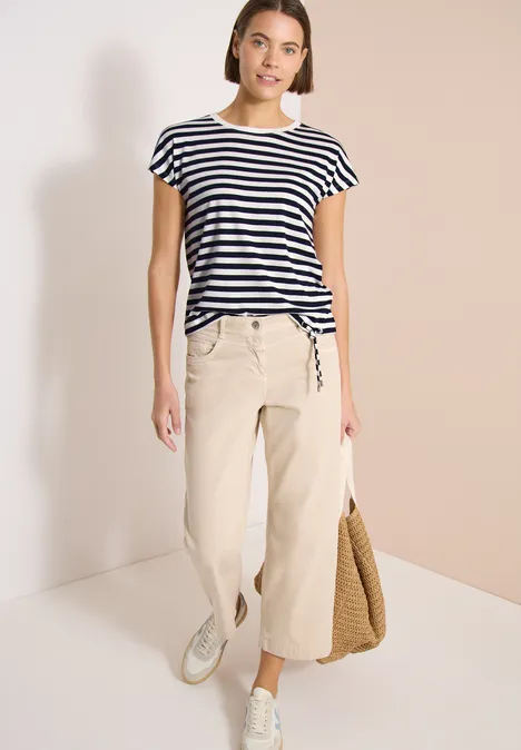 Femme en T-shirt rayé, pantacourt beige, baskets blanches, tenant un sac en paille.