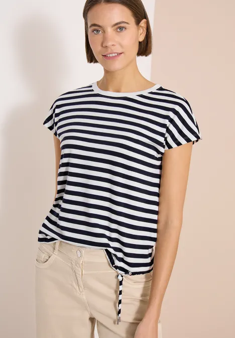 Femme en t-shirt rayé et pantalon beige devant un fond neutre.