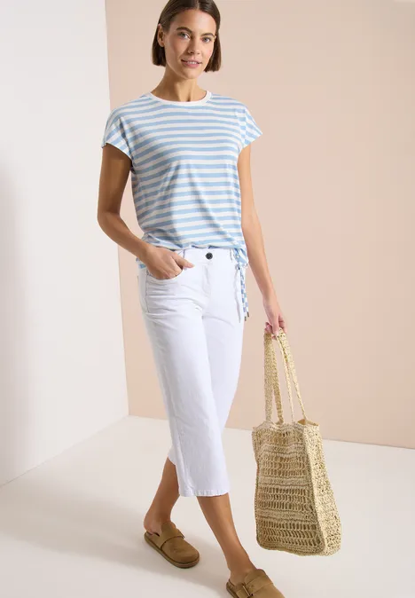 Frau trägt gestreiftes T-Shirt, weiße Capri-Jeans, Loafer-Schuhe und hält eine Korbtasche.