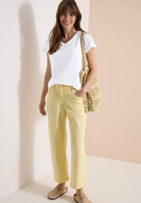 Frau in weißem T-Shirt und gelben Hosen, trägt Basttasche, vor hellem Hintergrund